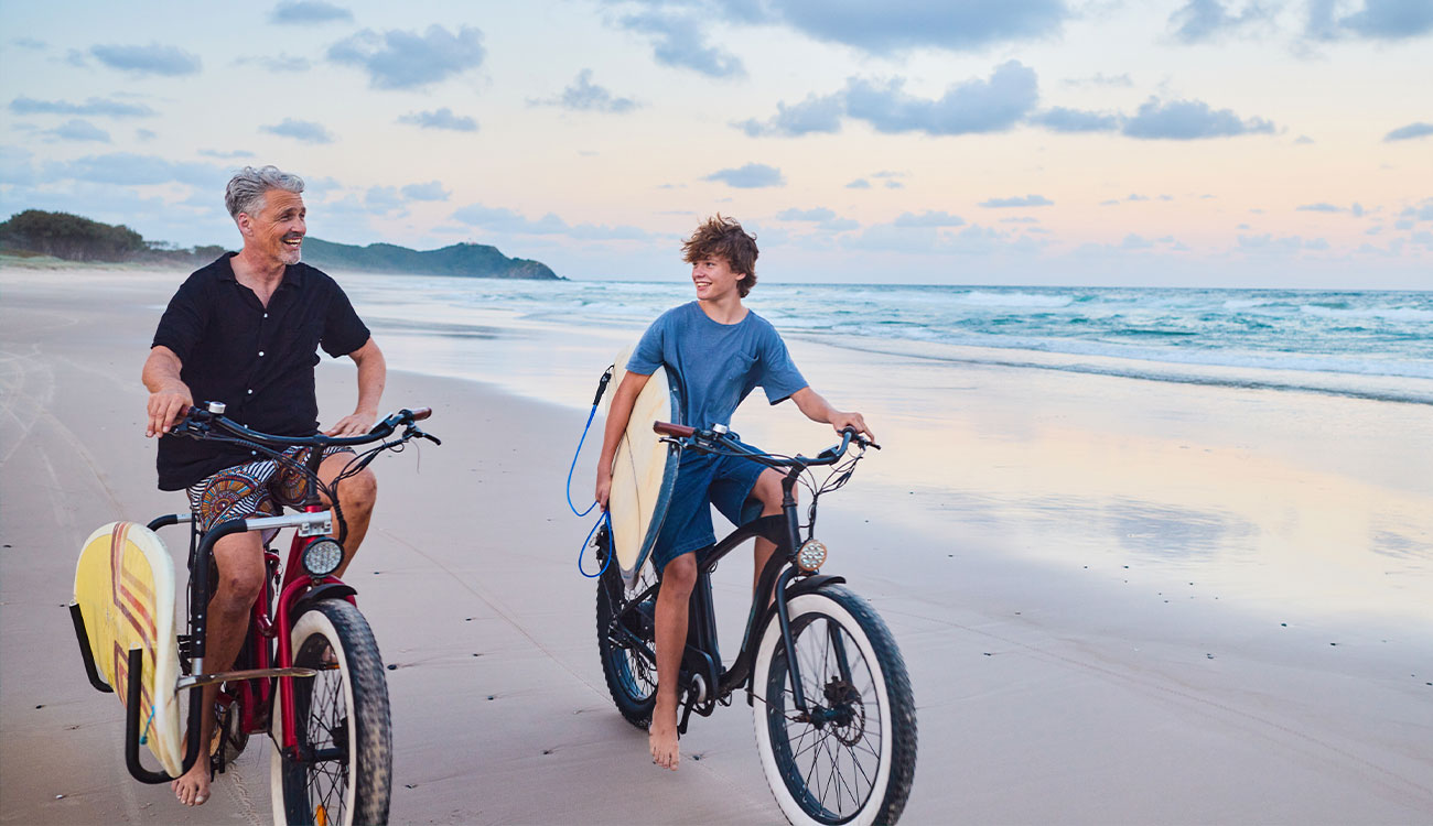Father and son riding bikes with surfboards on a peaceful beach at sunset.