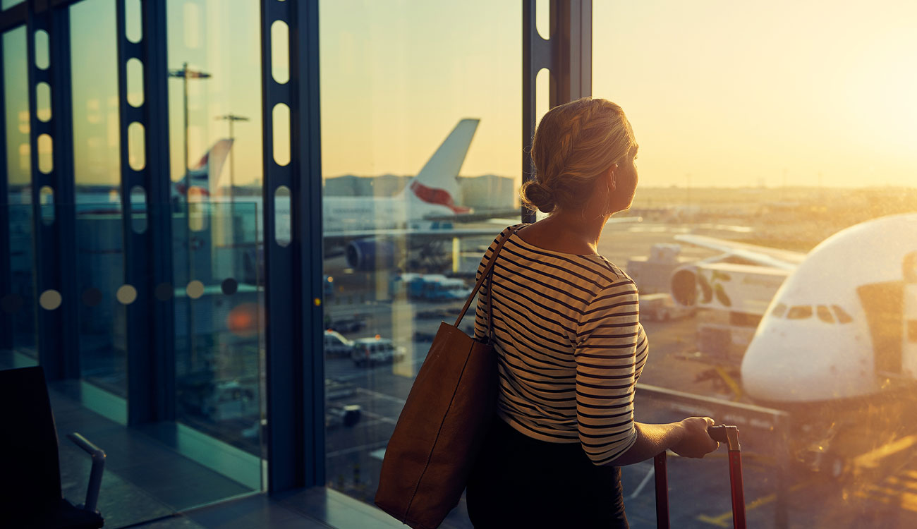 A woman stands inside an airport terminal, looking out through large windows at planes on the tarmac as the sun sets.