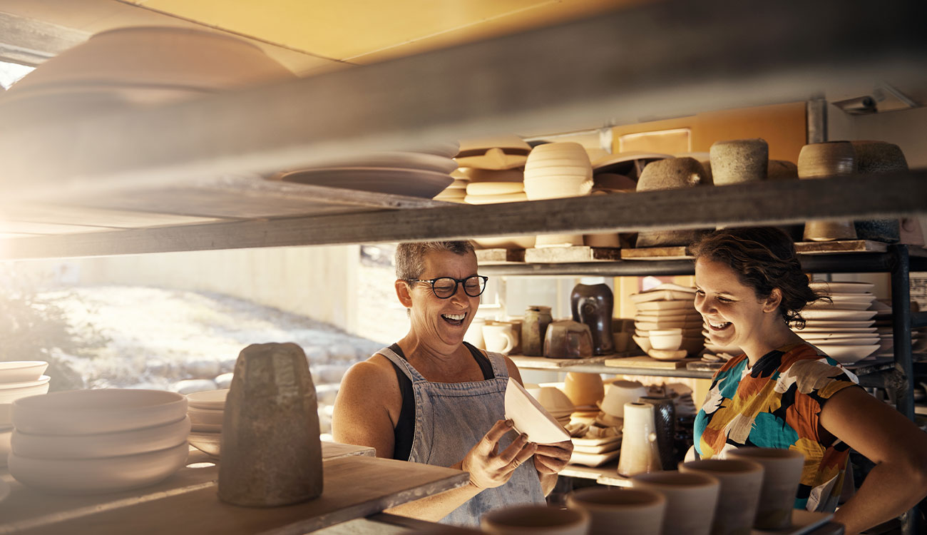 Two small business owners laughing together in a pottery studio, surrounded by handmade ceramics, representing local businesses supported through community-focused banking.