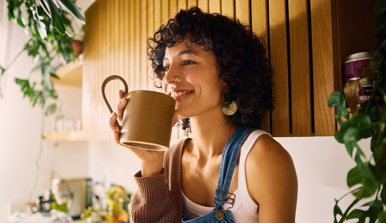Smiling woman drinking from a mug while relaxing in her kitchen surrounded by plants.