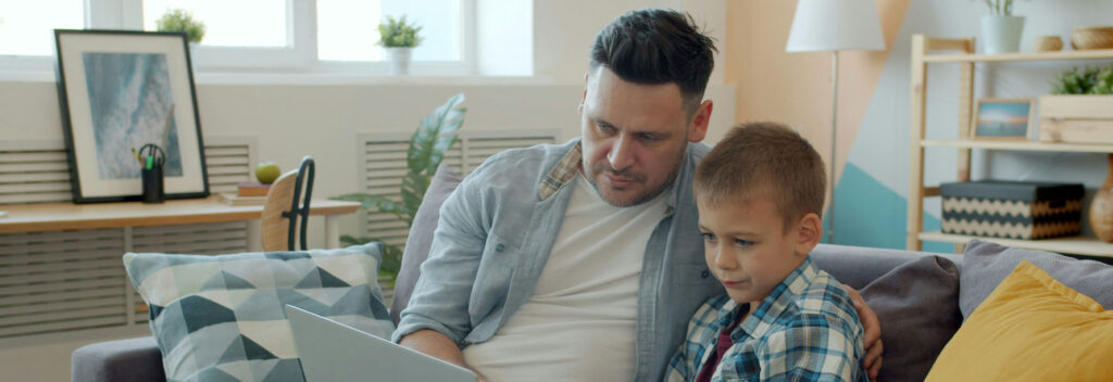 Parent sitting with child on a couch, helping them use a laptop safely at home.