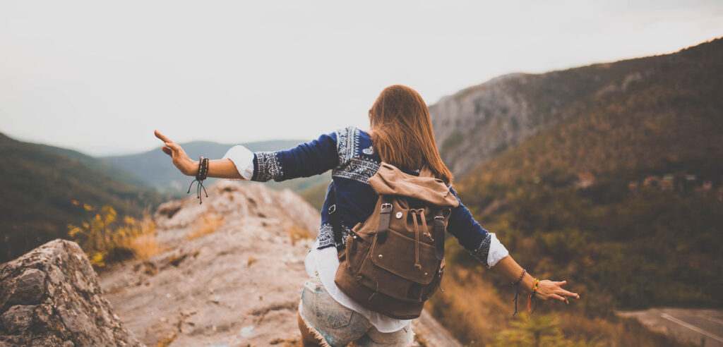Person wearing a backpack standing on a rocky lookout, arms outstretched, overlooking a wide valley and mountains.