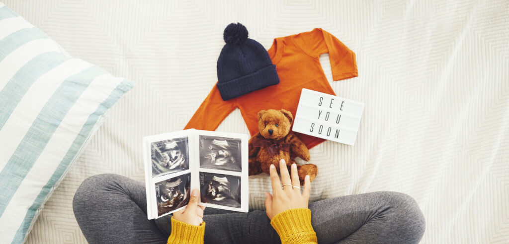 Ultrasound images held above baby clothes and a teddy bear laid out on a bed.