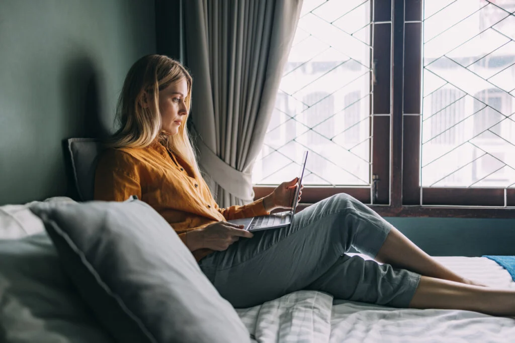 A Side View Of A Beautiful Blonde Female Working From Home On Her Laptop While Lying On The Bed Near The Window