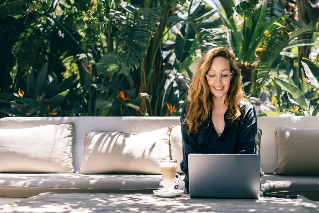 Young woman enjoying remote work in a tropical garden, sitting on a sofa with a laptop and latte. A peaceful and productive work-from-anywhere concept in a sunny and natural environment.